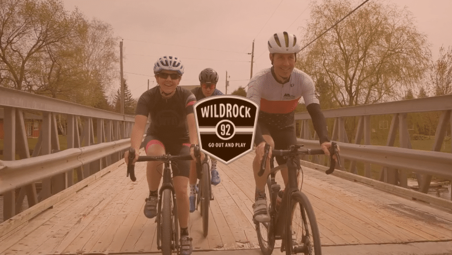 Cyclists cross a wooden bridge, with logo for outdoor company Wild Rock overtop