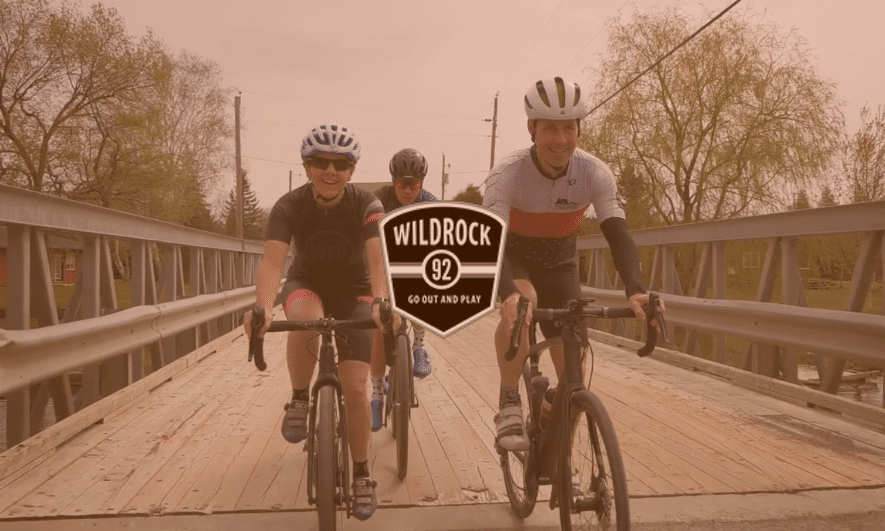 Cyclists cross a wooden bridge, with logo for outdoor company Wild Rock overtop