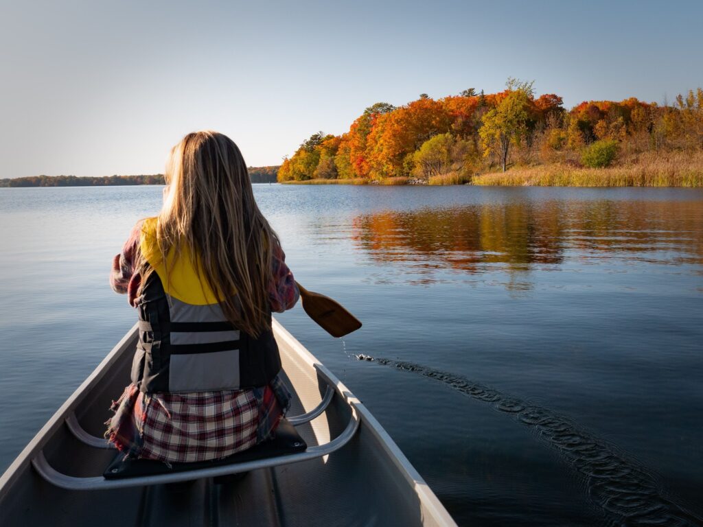 Woman paddling a canoe on a calm lake toward a shoreline of fall colours