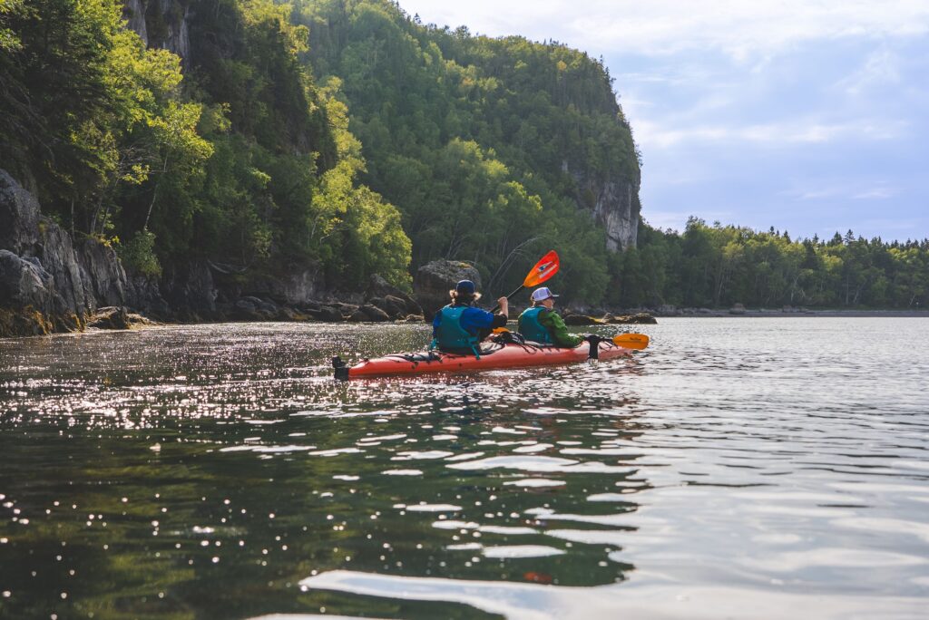 Two guests paddling a red tandem kayak beneath a forested cliff